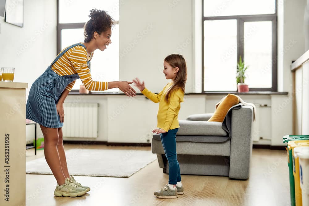 Nice to meet you. African american woman baby sitter get acquainted with caucasian cute little girl. They are standing indoors and talking